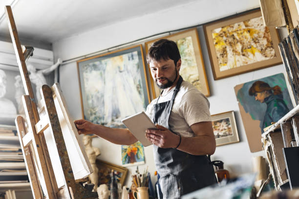 Male painter at art studio indoors copying picture from digital tablet on canvas concentrated