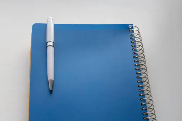 White pen attached to blue spiral notebook on white table. Selective focus. Background image with copy space for text
