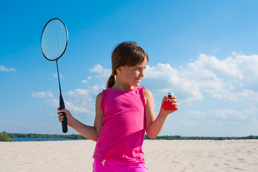 Outdoor portrait of a little girl with racket and shuttlecock in his hands by the river in Sunny day. Beach badminton, Concept sports and outdoor games in the summer.