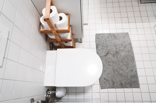 White tiles in a modern small bathroom, grey bath mat and bamboo toilet paper stand next to the toilet.