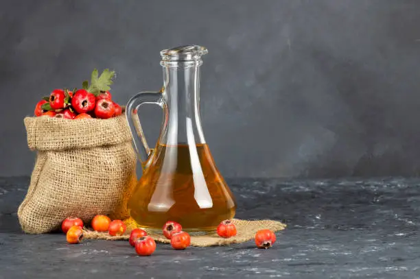 Glass bottle of hawthorn vinegar with Fresh ripe red/yellow hawthorn fruits on rustic background. Crataegus monogyna berries