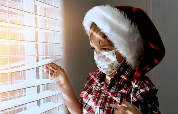 Little boy wearing a Santa hat and a protective face mask looking through a window waiting for Santa to came