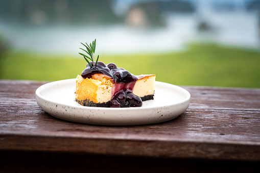 A piece of blueberry cheese cake served on wooden table-counter bar, with beautiful view of landmark bay location in Thailand as background. Dessert food close-up, selective focus photo.