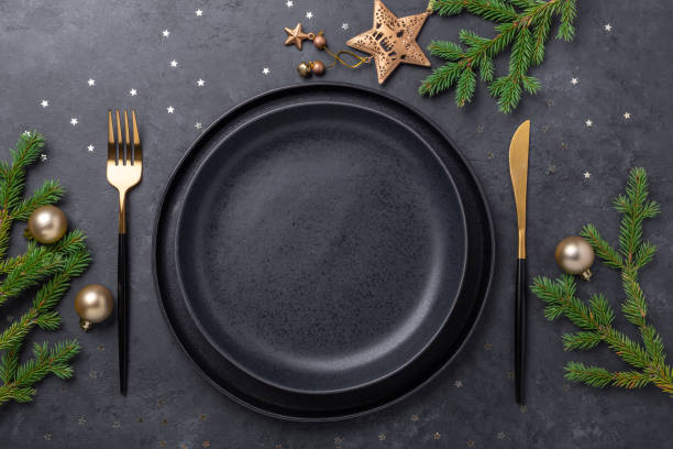 istock Christmas table setting with empty black ceramic plate, fir tree branch and gold accessories on black stone background. Top view. Copy space 1277482139
