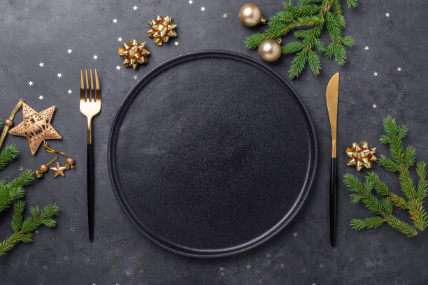 istock Christmas table setting with empty black ceramic plate, fir tree branch and gold accessories on black stone background. Top view. Copy space 1277482201