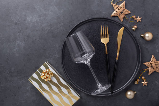 istock Christmas table setting with black ceramic plate, glass, gift box and gold accessories on black stone background. Top view. Copy space 1277482244