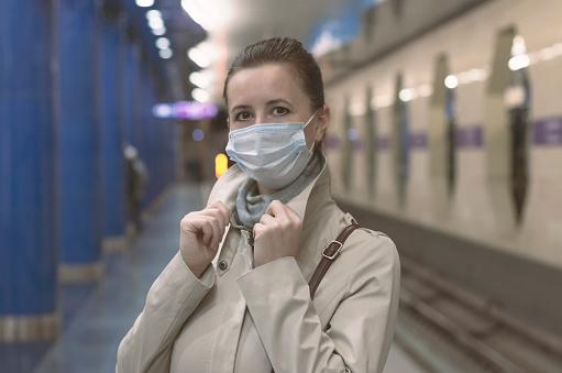 Young woman with a protective mask on her face on a deserted subway platform with bokeh lighting