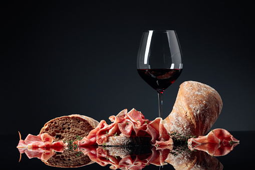 Prosciutto with ciabatta, red wine and thyme on a black reflective background. Copy space.