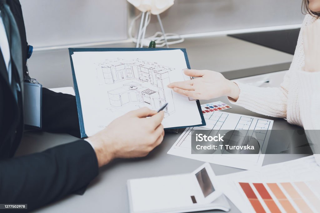 Seller shows kitchen layout and idea in a design. Close up manager in suit is showing blueprints to female client in kitchen store. The seller shows the kitchen layout and his idea in a design and flowers for her. Kitchen Stock Photo