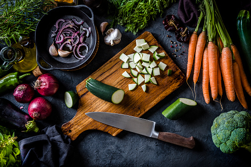 Top view of a wooden cutting board with chopped zucchini on top surrounded by vegan ingredients like carrots, onion, broccoli, eggplants, basil, garlic, olive oil, rosemary and a bell pepper. The cutting board is at the center of the image and it is on a grey kitchen tabletop. Low key DSLR photo taken with Canon EOS 6D Mark II and Canon EF 24-105 mm f/4L