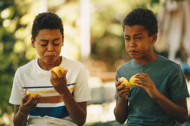 deux frères d’origine africaine s’asseyant à l’extérieur et goûtant le citron. - grimacing male african descent teenage boys photos et images de collection