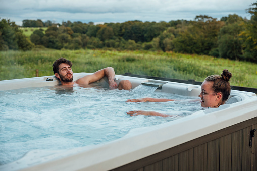 A shot of a mid adult man and young woman sitting in a hot tube in a rural setting.