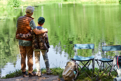 Full length back view portrait of father and son standing by lake and enjoying nature during hiking or fishing trip, copy space