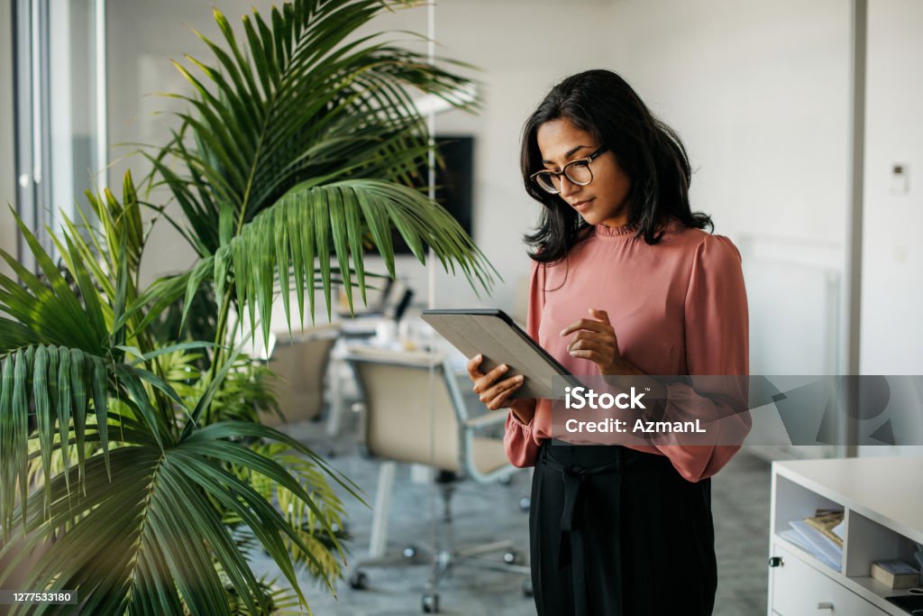 Young Indian Businesswoman Using Digital Tablet in Office Female executive with medium-length black hair wearing eyeglasses and businesswear standing outside modern meeting room and checking data on tablet. Office Stock Photo