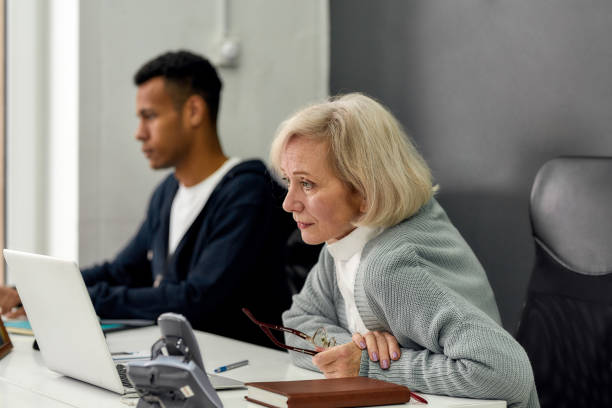 istock Aged woman, senior intern looking focused at the screen while using laptop, sitting at desk, working in modern office with other young employee 1277535025