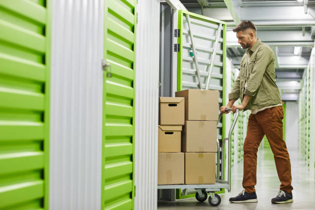 Man Moving Boxes in Storage Unit Full length side view at handsome bearded man loading cart with cardboard boxes into self storage unit, copy space storage compartment stock pictures, royalty-free photos & images