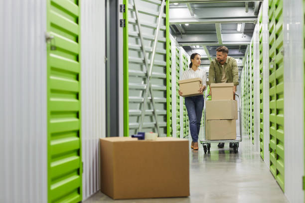 Couple Moving Boxes in Storage Unit Full length portrait of young couple holding cardboard boxes walking towards camera in self storage unit, copy space storage compartment stock pictures, royalty-free photos & images