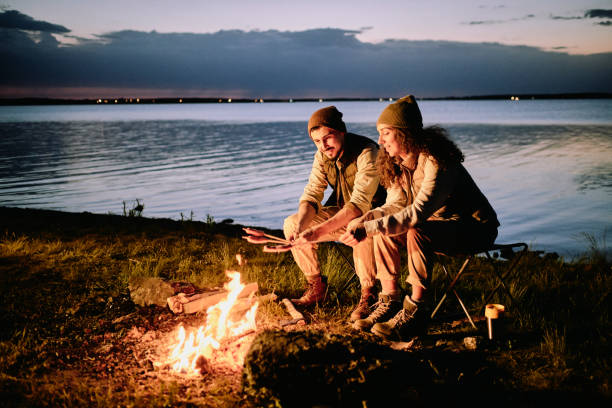 Making sausages on fire during camping Positive young couple sitting at lake shore and making sausages on fire during night camping Bonfire stock pictures, royalty-free photos & images