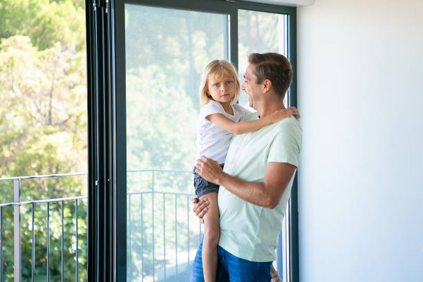 Cheerful father holding cute daughter, looking at her and smiling. Lovely blonde girl looking at camera. Happy dad with kid standing near balcony open door. Relocation and family moving day concept