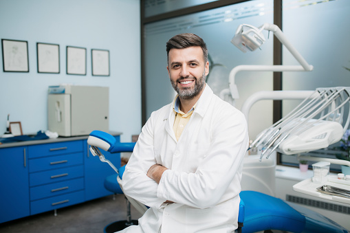Portrait of a Caucasian man dentist, sitting in his office next to a dentist chair, smiling