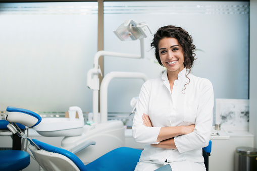 Portrait of a Caucasian woman dentist, sitting in her office next to a dentist chair, smiling