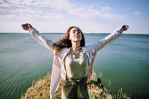 Free attractive curly-haired girl in casual shirt standing with crossed eyes and outstretched arms and enjoying wind at seaside