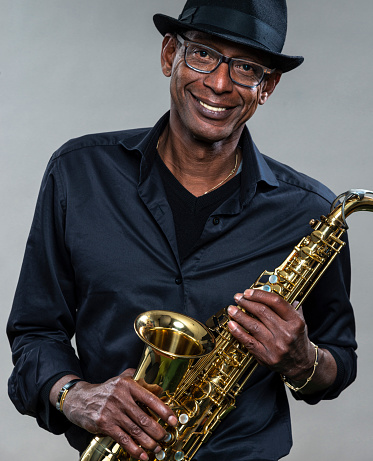 Musician with saxophone  against a grey backdrop. Man looking at camera with a smile, wearing a black hat and shirt
