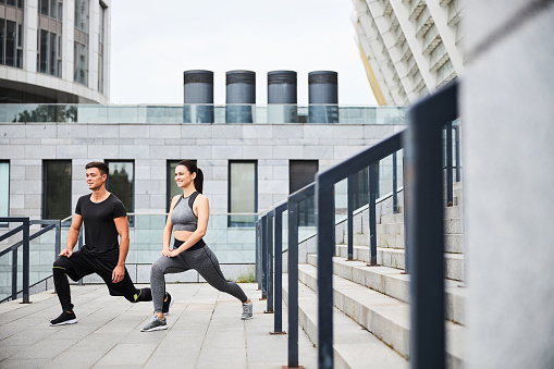 Happy slim woman and man are doing lunges while training on stairs to urban stadium