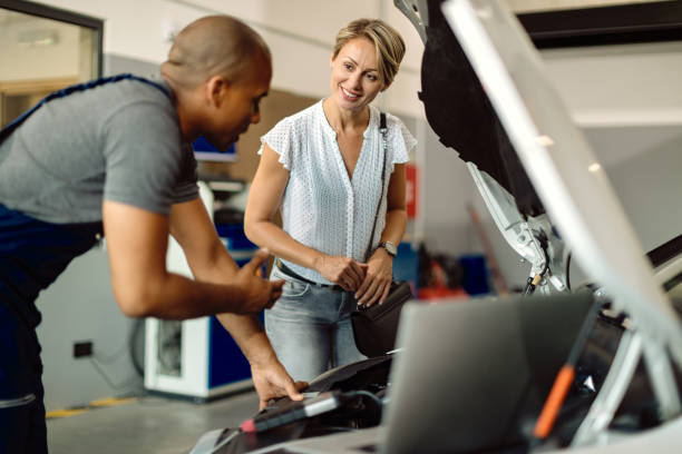 Happy woman talking to her car mechanic in a repair shop. Young happy woman communicating with her auto repairman who is examining engine problems in a workshop. auto repair shop stock pictures, royalty-free photos & images