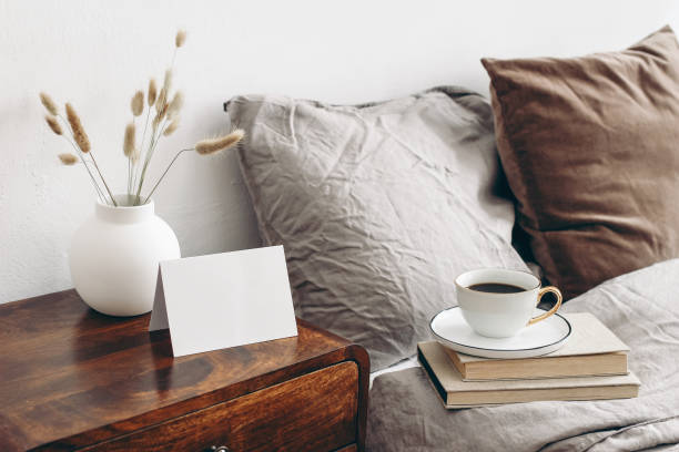 istock Empty greeting, place card mockup on retro wooden bedside table. Modern white ceramic vase with dry Lagurus ovatus grass and cup of coffee. Beige linen and velvet pillows. Scandinavian bedroom interior. 1277563940