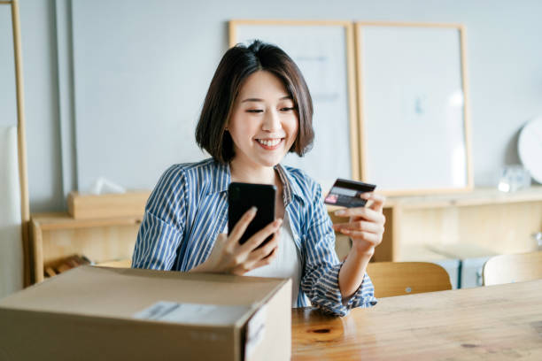 jeune femme asiatique gaie faisant des achats en ligne avec le smartphone et faisant le paiement mobile avec la carte de crédit à portée de main, recevant un colis par service de livraison à domicile. la technologie rend la vie tellement plus facile - qr code marketing mobile phone coding photos et images de collection