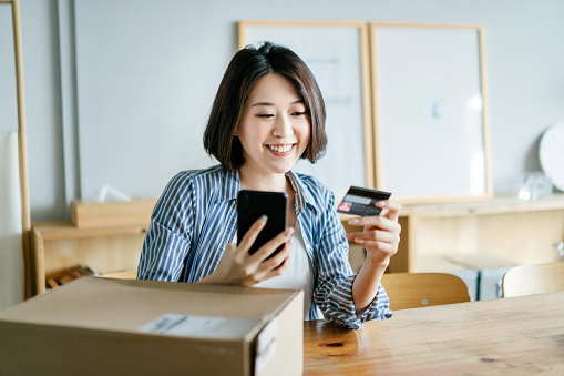 Cheerful young Asian woman shopping online with smartphone and making mobile payment with credit card on hand, receiving a parcel by home delivery service. Technology makes life so much easier