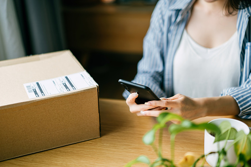 Close up of young Asian woman preparing a parcel for delivery using mobile app device on smartphone at home. Technology makes life so much easier