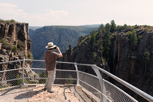 Rearview photograph of a Caucasian man wearing a hat while taking photos at a scenic viewpoint in Black Canyon of the Gunnison National Park in Colorado, USA on a sunny summer day.