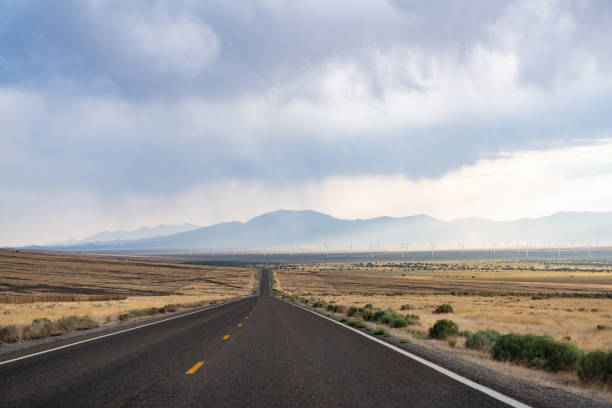 This is a wide angle photograph of empty rural highway 50 through the Nevada desert landscape on a cloudy day. Windmills can be seen far in the distance.