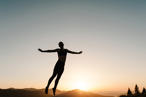 Happy woman jumping and enjoying life at sunset in mountains. Girl doing fitness exercise sport outdoors in the morning. Healthy lifestyle concept. Freedom, risk, challenge, success.