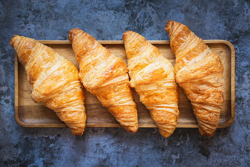 Four freshly baked French croissants on a dark wooden tray. The background underneath them is a dark.