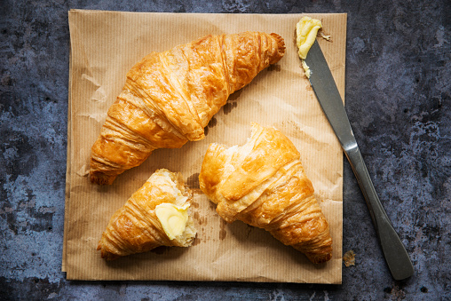 Two freshly baked French croissants are sitting on a brown paper baker’s bag. One has been torn in half and some butter has been smothered across one end. There is a knife with butter on it resting on the bag beside the croissants. The background underneath them is a dark.