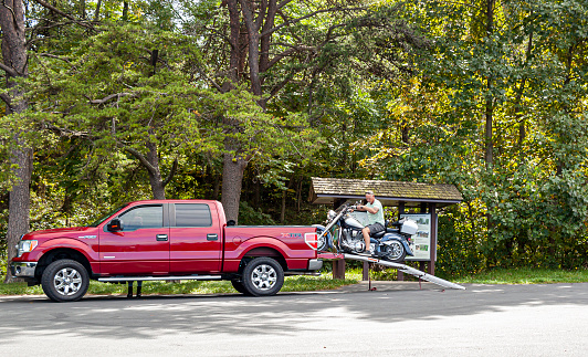 Shenandoah Valley, VA, USA 09/27/2020: An elderly motorist is unloading his motorbike from the trunk of a red Ford F150 XTR pickup by slowly backing up on a metal makeshift ramp at Shenandoah Park.