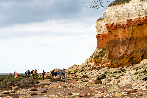 Hunstanton, Norfolk, East Anglia, England, UK. The chalk and sandstone cliffs are constantly being eroded and collapsing on to the beach.  This shows the different coloured rock strata and a pile of collapsed rocks on the beach. There are crowds of people on the beach and some perilously close to the unstable cliffs.