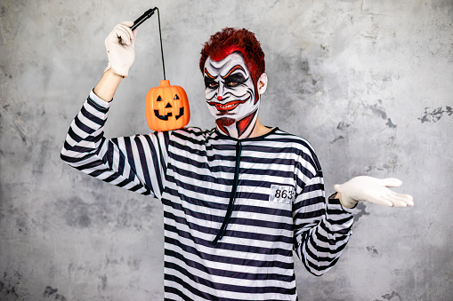Mid adult man in prisoner costume and painted face imitating a killer clown while holding a toy pumpkin during halloween season