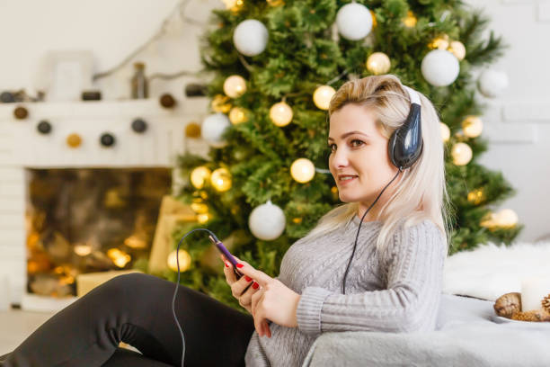 femme heureuse appréciant sa musique à noël se relaxant sur un sofa devant l’arbre avec un sourire rayonnant serrant son portable à sa poitrine. - tree house audio photos et images de collection