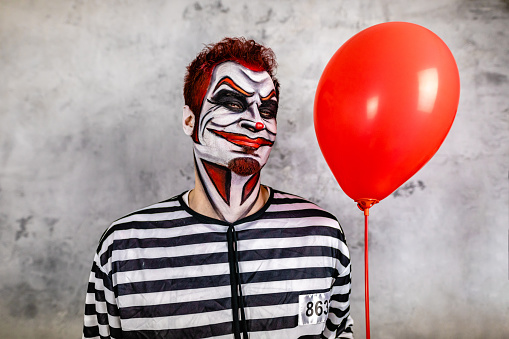 Mid adult man in prisoner costume with red hair and red contact lens imitating a scary  angry clown while holding a red balloon for a Halloween event