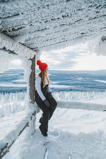 Young traveler woman wearing winter clothes and warm gloves enjoying  beautiful sunset view while sitting in a wooden hut in a breathtaking deepsnow covered forests and mountain ranges snowy winter landscape in