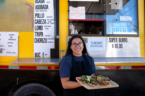 Pretty Young Mexican Woman in front of her small business food truck taco stand smiling at the camera and displaying a variety of her delicious gourmet tacos
