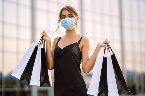 Young woman in protective sterile medical mask on her face with shopping bags near shopping center. Shopping during the coronavirus Covid-19 pandemic. Purchases, black friday, discounts, sale concept.