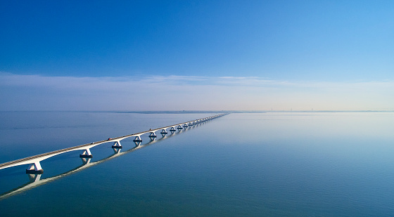 The Zeelandbrug during sunrise. The Sky is blue and it’s reflected in the water of the North sea. The bridge is also reflected in the water. Cars drive at a high speed over it.