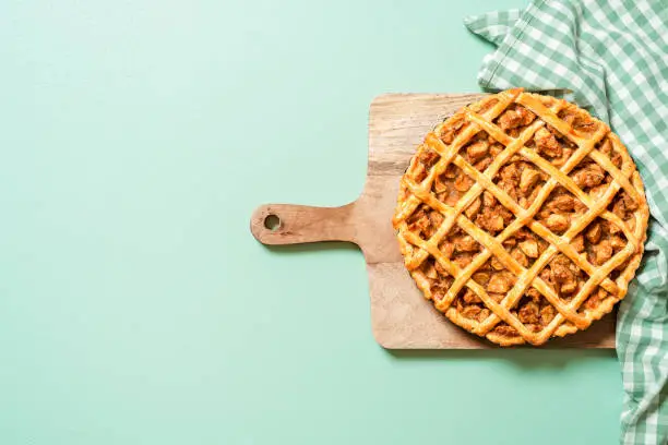 Above view with a freshly baked apple pie on a green colored table, home-baked apple pie with a lattice crust on a wooden board.