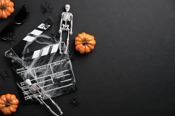 Photo of Halloween scary movie concept. Flat lay composition with clapper board, skeleton, spider web, pumpkins on black desk. Halloween background.