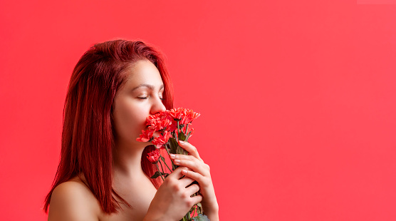 Young woman with beautiful skin, red hair, and closed eyes in front of a colorful background. Woman holding with both hands a bunch of red flowers.